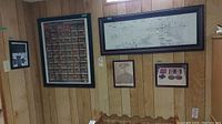 Wide shot of wood-paneled wall displaying a large framed Avro Arrow blueprint at top center, a Canada Victoria Cross chart poster on left, and smaller framed portrait and medal images below.