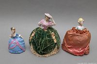 Overview of three dolls: two half porcelain doll pin cushions with fabric skirts and one porcelain trinket box doll in blue dress with pink trim.