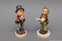 Front view of two Goebel porcelain figurines of boys, one playing an instrument, the other holding sheet music.