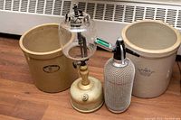 Full lot including two stoneware crocks, gas lamp, and seltzer bottle on wood floor in front of heater.