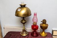 Three old hurricane oil lamps on a cloth-covered surface, including large converted brass lamp without chimney, red glass lamp with chimney, and a brass lamp.