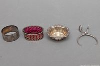 Four sterling silver pieces shown on a gray surface, including napkin ring, cranberry red glass lined item, salt cellar, and sugar tongs.