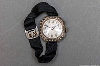 Antique wristwatch front view showing octagonal diamond-set bezel and white dial with black numerals and blue hands on a black ribbon strap.