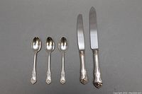 Top view of three sterling silver handled spoons and two knives with sterling silver handles and stainless steel blades, laid flat on a gray surface.