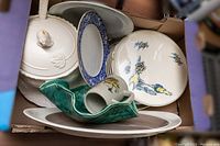 Various dishware items inside box including white tureen, blue floral bordered plates, round plates with floral and bird designs, green leaf-shaped dish, small pitcher with floral details.