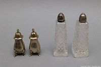 Four salt and pepper shakers arranged side by side, two small sterling silver shakers with lids on the left and two tall cut crystal shakers with sterling silver lids on the right.