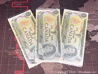 Three Canadian one dollar banknotes laid out side by side showing front arabesque and portrait of Queen Elizabeth II.