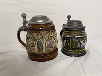 Two vintage German beer steins side by side, showing the difference in size and detailed colorful relief designs.