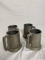 Four grey metal steins with broad handles and stepped bases on a white background, showing varying angles and grouping.