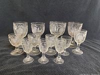 Front view of twelve assorted EAPG goblets and glasses arranged on black fabric background, showing variations in size and pattern details.