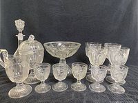 Group photo showing the entire set: candy dish with lid, pedestal dish, decanter, assorted goblets, and a small vase. All pieces have clear pressed glass with grape pattern.