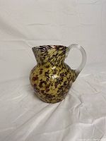 Front and side view of hand blown glass pitcher showing rounded, mottled patterned body, clear handle, and spout.