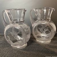 Side view of two clear glass pitchers with round embossed fern patterns and textured handles