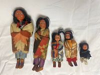 Five Native American dolls of various sizes on white fabric background, showing full body and clothing details.