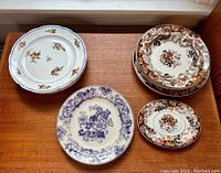 Seven antique plates arranged on a wooden surface showing varying floral and ornamental patterns in colors including white with colorful flowers, brown-orange ornamental pattern, and purple ornamental design. Some plates display chips and cracks visible to the camera.