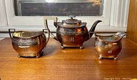 Full set of three silver plated tea set items on wooden surface in front of window: teapot, creamer, sugar bowl, each with engraving and letter M monogram.