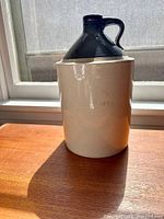 Full side view photo of the antique stoneware jug showing its two-tone coloration with a dark neck and handle and lighter body. The jug stands on a flat surface with natural light.