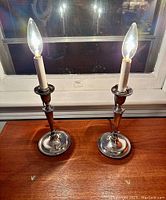 Pair of antique sterling silver candlestick lamps lit and placed on a wooden surface near window at night showing reflective surfaces.