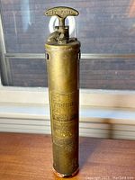 Full view of vintage brass fire extinguisher on wooden surface with window background showing pump handle and body.