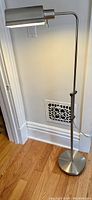Full view of the chrome floor lamp standing on wooden floor against a wall, showing base, pole and swing arm with shade.