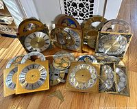Photo showing a selection of grandfather clock faces laid out on wood floor, including multiple square brass-colored faces with Roman and Arabic numerals, circular moon phase dials, and some loose parts.