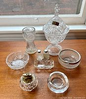 Front view of all crystal pieces on wooden surface, showing candy dish with lid, vase, salt and pepper shakers, trinket dishes and lighter