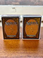Two stacks of Magic the Gathering cards shown from the back with protective cases on a wooden surface, displaying the iconic card back design.