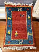 Full top view of the folk carpet mat with colorful animal and human figures along the border and central red field.