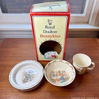 Set consisting of a Royal Doulton Bunnykins box, Bunnykins mug, Bunnykins bowl, and Wedgwood Peter Rabbit bowl arranged on a wooden surface in front of a window.