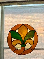 Photo showing hanging stained glass piece with orange circular border and fleur symbol in amber and green color glass in front of a window.
