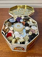 Photo showing open decorative tin filled with various buttons of different colors, sizes, and materials including plastic and metal.