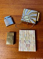 Photo showing all four items on a wooden table: Ronson lighter, gold briquet lighter, sunburst enamel compact/cigarette case with chain, and mother of pearl cigarette case.