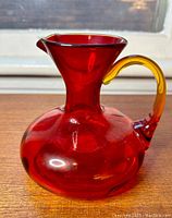 Profile view of the red glass pitcher showing rounded base, flared neck, and amber handle attached at the back.