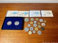 Photo shows a collection of twenty-six coins including four in protective holders and two in a blue velvet-lined box, arranged on a wooden surface.
