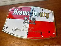 Front view of vintage Kronenbourg metal wall clock on wood surface, showing red and white background with logo and clock hands at 12:00.
