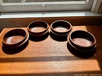 Four vintage Danish teak bowls arranged in a row on a wooden surface with sunlight illuminating them, showing round shape and warm teak color.