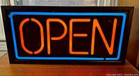 Front view of vintage neon open sign illuminated, showing bright red-orange 'OPEN' with a blue border in black frame.
