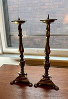 Two vintage brass candlesticks standing on a wooden surface by a window, showing full height and form.