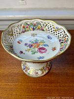 Angled front view of the porcelain compote showing floral pattern inside, reticulated border, and gold rim.