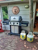 Front view of Broil King BBQ grill with two propane tanks positioned in front and side shelves visible.