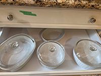 Three covered CorningWare FrenchWhite stoneware casserole dishes shown from above inside a kitchen drawer, displaying one smaller and two larger dishes with clear glass lids.