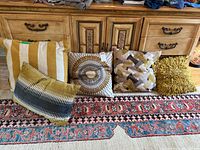Five decorative throw pillows arranged on floor in front of wooden dresser, showing various sizes and patterns.