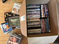 Photo showing a large cardboard box filled with assorted DVD movie cases stacked upright. Several other stacks of DVD and CD cases lie beside the box on the floor.