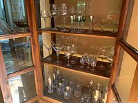 Three shelves of assorted clear glassware including wine glasses, martini glasses, and liquor glasses arranged neatly inside wooden cabinet.