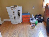 Photo showing white plastic laundry hamper, small organizer cabinet, DVDs, digital clock radio, papers, and miscellaneous items on hardwood floor.