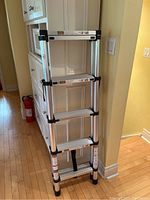 Folding telescopic aluminum ladder in collapsed storage position against white cabinetry and hardwood floor.