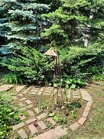 Full view of taller metal candle holder standing in garden bed with brick edging, rusted with butterfly cutout design on shade.