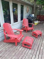 Two red painted Muskoka chairs and two matching footstools shown outdoors on a wooden deck next to a house.