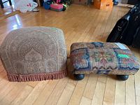 Two cushioned ottomans side by side on wooden floor. One with beige paisley fabric and fringe, the other rectangular with floral tapestry fabric and black ball legs.