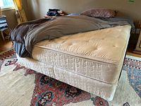 Photo showing queen size mattress with beige tufted surface and box spring underneath on a colorful patterned rug in a room with curtains and furniture.